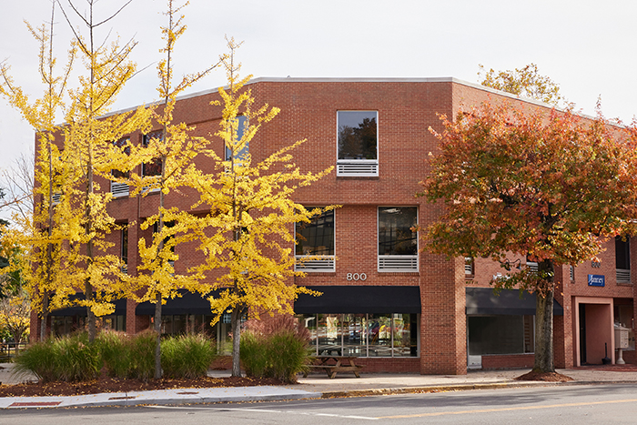 The image shows a two-story brick building with a flat roof, situated on a street corner. In front of the building is a tree with yellow leaves, indicating autumn season, and the sidewalk is lined with trees and shrubs.