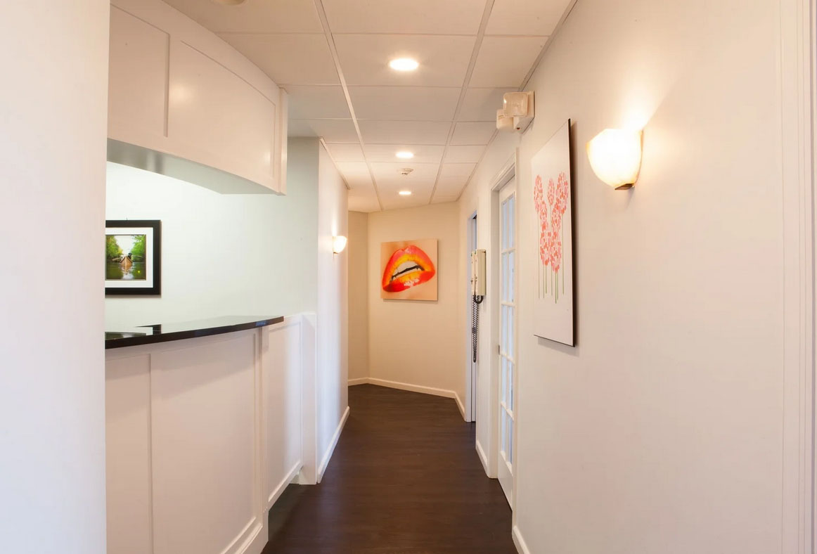 The image shows an interior hallway with white walls, a dark wood floor, a kitchenette on the left with white cabinets, a black countertop, and a refrigerator, and artwork hanging on the wall. There are two doors at the end of the hall, one on each side, leading to separate rooms. The lighting is bright, and the overall style of the image suggests it may be a real estate photograph intended for promotional purposes.