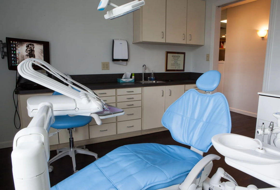 The image shows an interior view of a dental office with modern equipment, including a dental chair, sink, and a large screen displaying a patient s face.