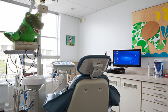 The image shows an interior view of a dental office with a chair, desk, and other medical equipment, featuring a dinosaur toy on a shelf and a cartoon character poster on the wall.