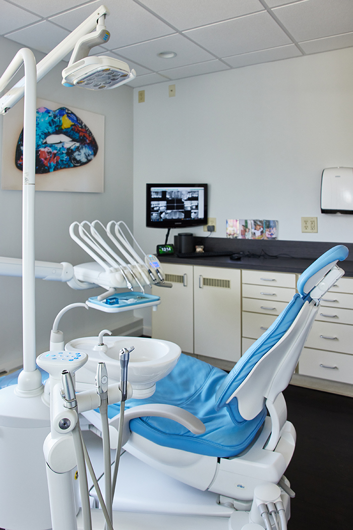 The image shows a well-equipped dental office with a modern chair, open cabinets, and a television screen displaying an advertisement for dental services.