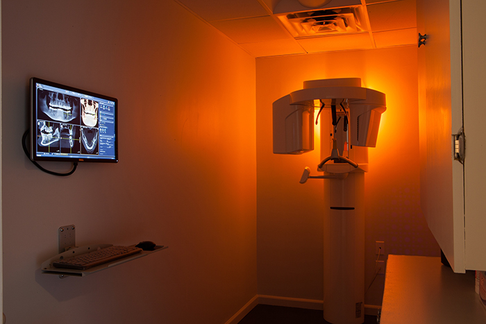 The image shows an interior room with medical equipment, including a dental chair and a large screen displaying an X-ray.