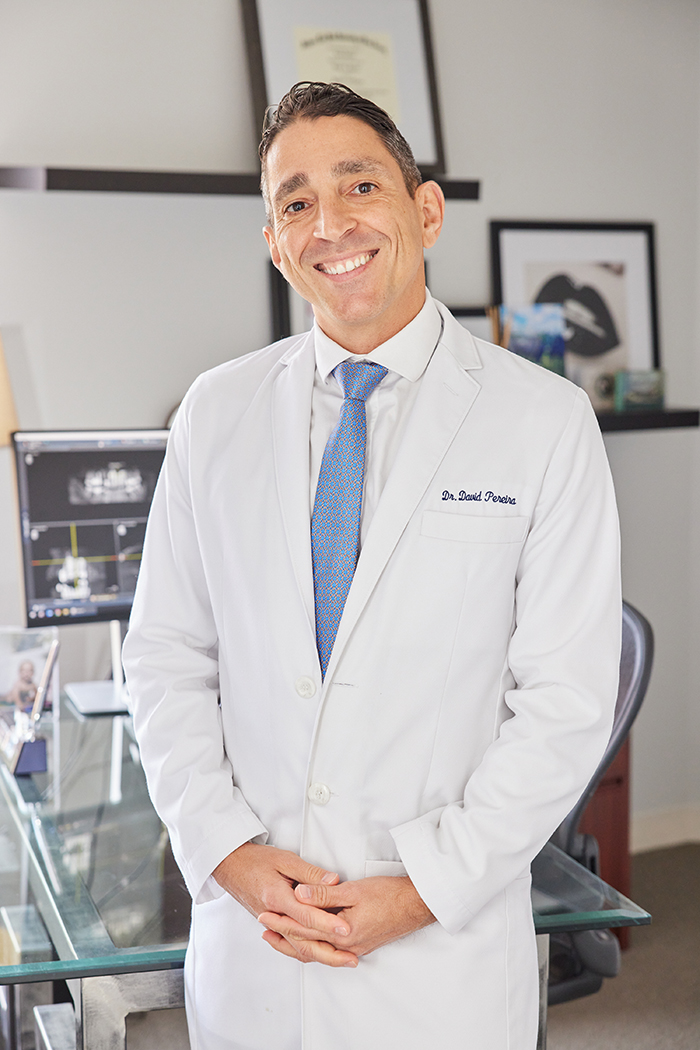 The image shows a man wearing a white lab coat standing behind a desk with a smile on his face, posing for a portrait photograph.