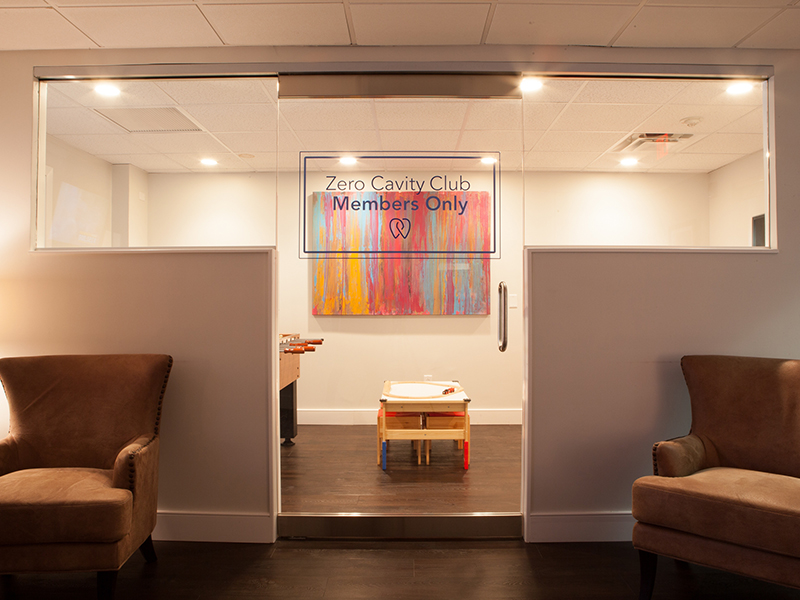 The image shows an interior space with a sign indicating 'Zero Cavity Club Members Only.' There are two couches, one visible through the glass door on the left and another partially visible on the right side of the frame. The room has a modern aesthetic with neutral colors and a large window.