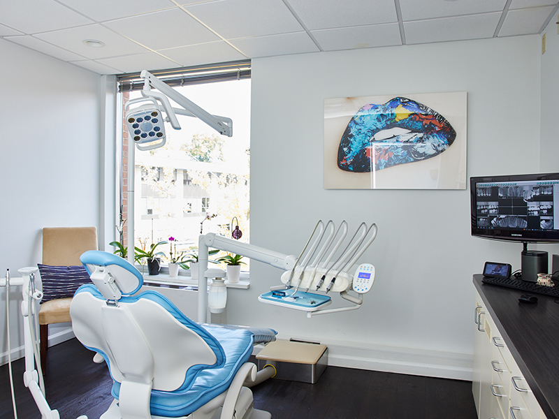 The image shows a dental office interior with a modern dental chair, a large screen mounted on the wall displaying an X-ray image, and other dental equipment visible in the background.