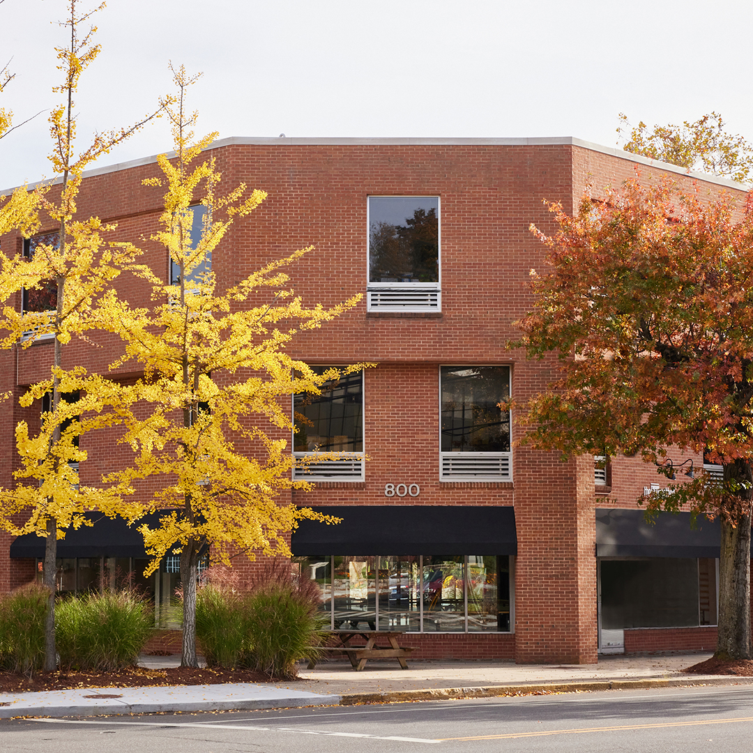 The image shows a two-story brick building with a flat roof, featuring large windows and a black canopy over the entrance. In front of the building, there are trees with yellow leaves, indicating it might be autumn. There s a sign on the building that reads  800,  suggesting it is a street address or a suite number. The building is situated at an intersection, as indicated by the presence of a road and sidewalk in the foreground.