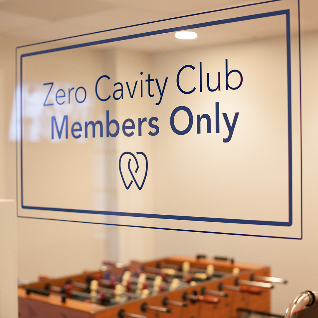 The image shows a sign indicating  Zero Cavity Club  membership only for zero cavities, with a background featuring a foosball table.