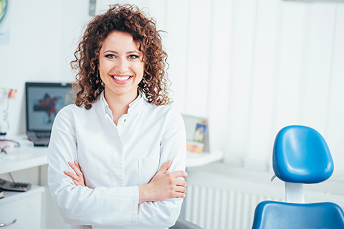 A woman with curly hair stands confidently in a dental office, smiling at the camera.
