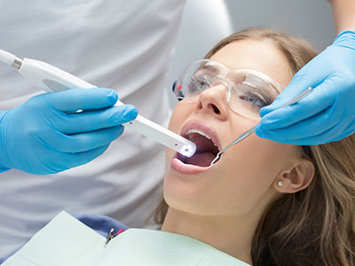 A woman receiving dental care with a device inserted into her mouth while wearing protective blue gloves and glasses.
